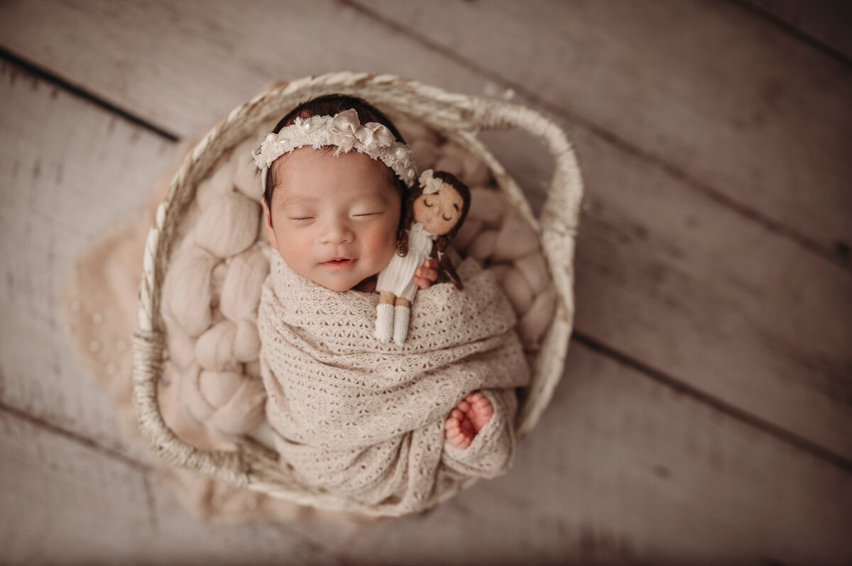 Smiling newborn baby girl  holding doll lovey while wrapped in a cream colored wrap and wearing matching headband laying inside basket prop over a chunky braided blanket with a wooden  floor background