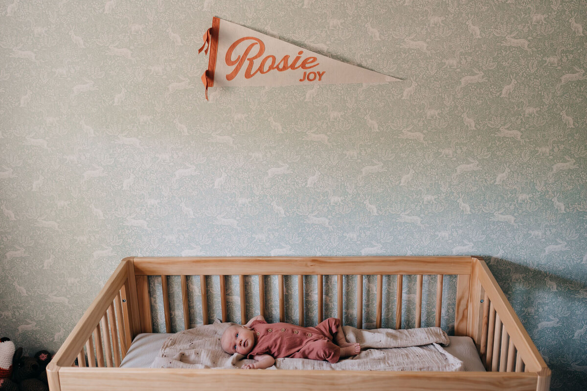 A baby lays awake in her crib in her nursery, a pendant with her name is on the wall and the wall has soft green woodland wallpapaer