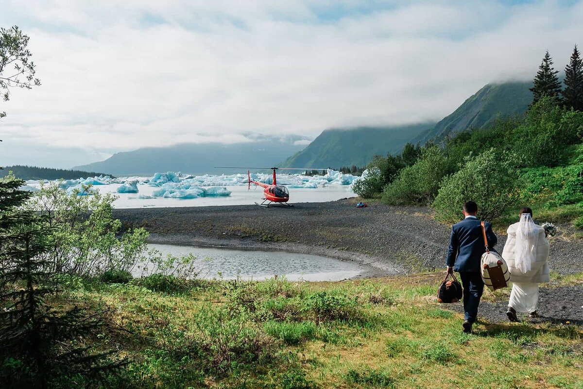 The bride and groom are dressed in their wedding finest, walking across a dark pebbly beach toward the bright red private helicopter that has come to pick them up during their  two day Bear Lake luxury elopement with dogsledding in Alaska. Behind the heli, the background is innumerable blue-tinted white icebergs floating in the beautiful turquoise water of and rugged snow capped mountains in the distance, shrouded in low clouds.   The groom carries their classic luxury styled, leather and canvas overnight luggage satchels. 