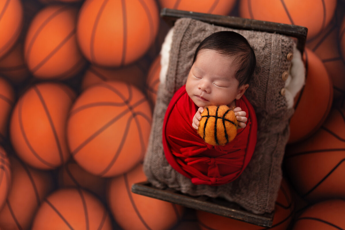 Basketball-themed newborn photo of baby boy wrapped in red and holding mini basketball on sports backdrop.