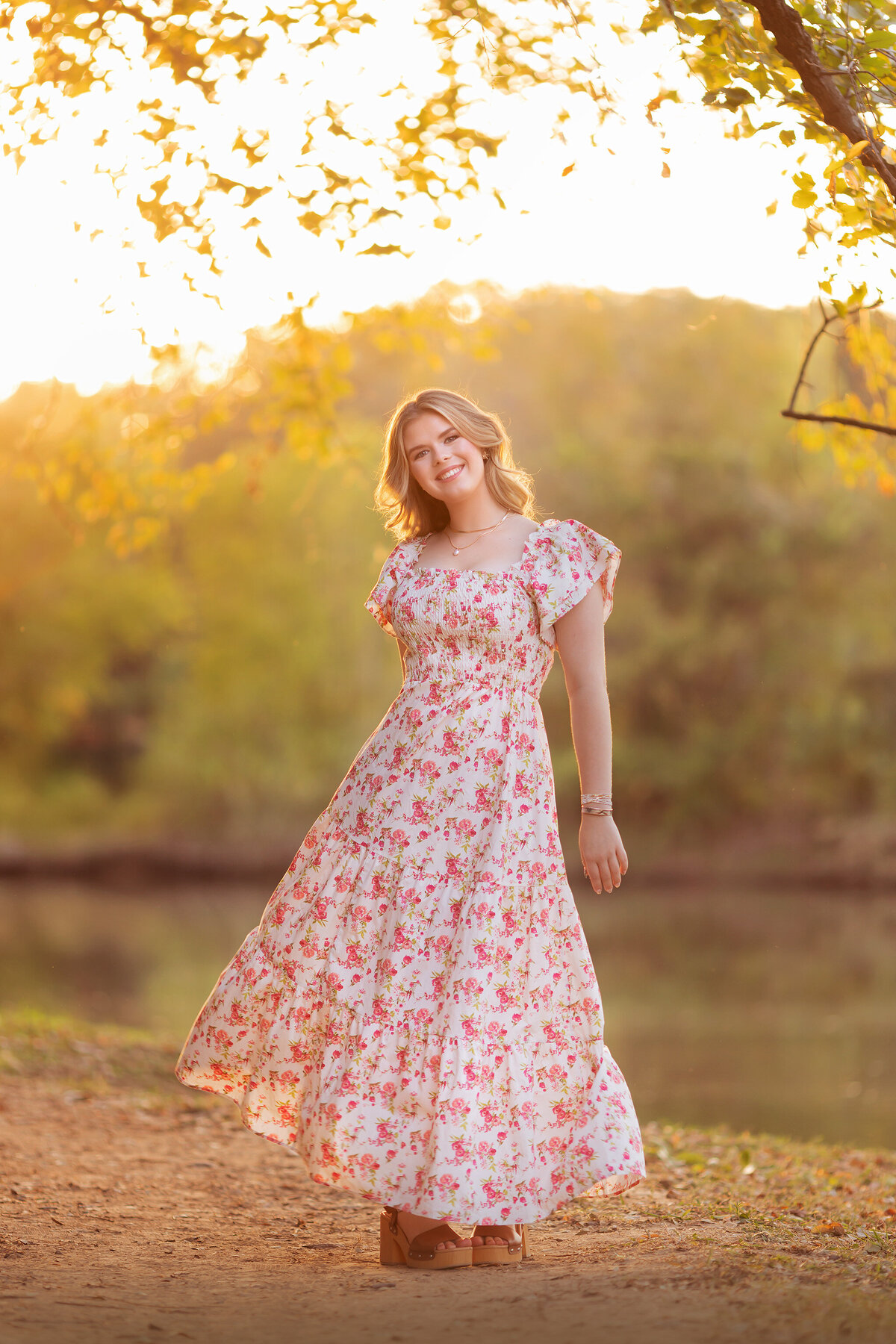 Dallas senior photographer captures a young woman in a floral dress, twirling in golden light by a serene river.