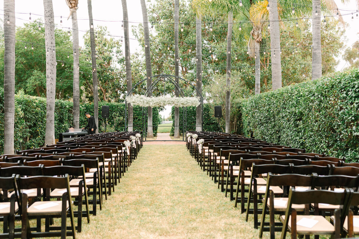Outdoor wedding setup with two rows of empty black chairs on a grassy aisle. Greenery-filled arch and string lights create a serene, elegant ambiance.