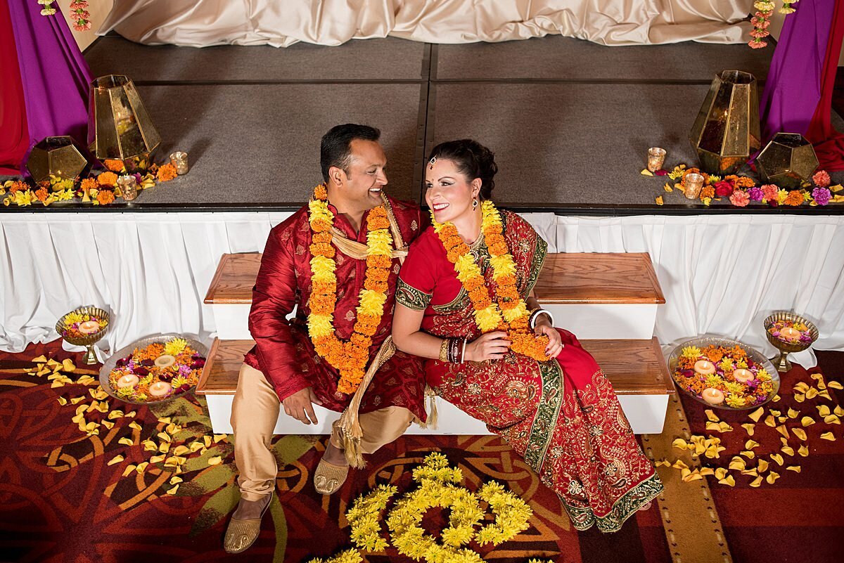 Indian Groom and bride sitting at the edge of the mandap wearing orange and yellow varmala garlands laughing.