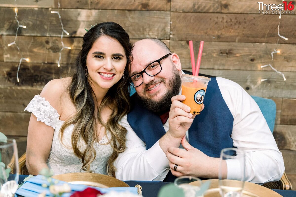 Bride and Groom pose together sitting at their sweetheart table holding a cup of Boba Drink
