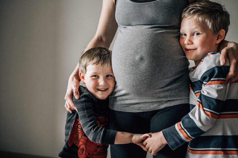 Two young boys holds hands with each other and hug their mother's growing belly during an in home maternity photography photoshoot.