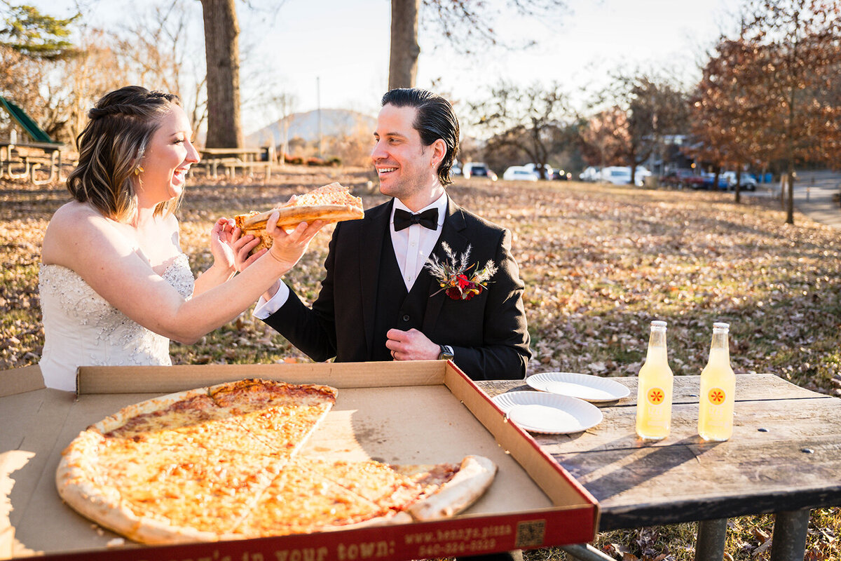 A couple on their elopement day intertwine hands and feed each other pizza at a picnic table at Mill Mountain in Roanoke, Virginia. The table features a large pizza from Benny Marconi’s and two IZZE drinks.