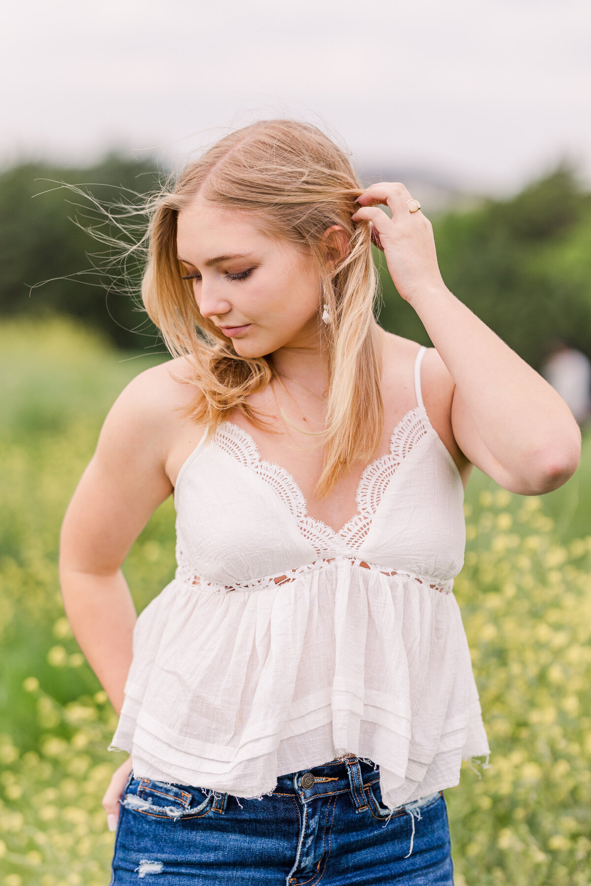Close-up senior portrait of a young woman with blonde hair in a white lace-trimmed top, gently brushing her hair back while standing in a sunny green field at Arbor Hills Nature Preserve in Plano, TX. A light and airy senior photo with natural beauty and soft expression.