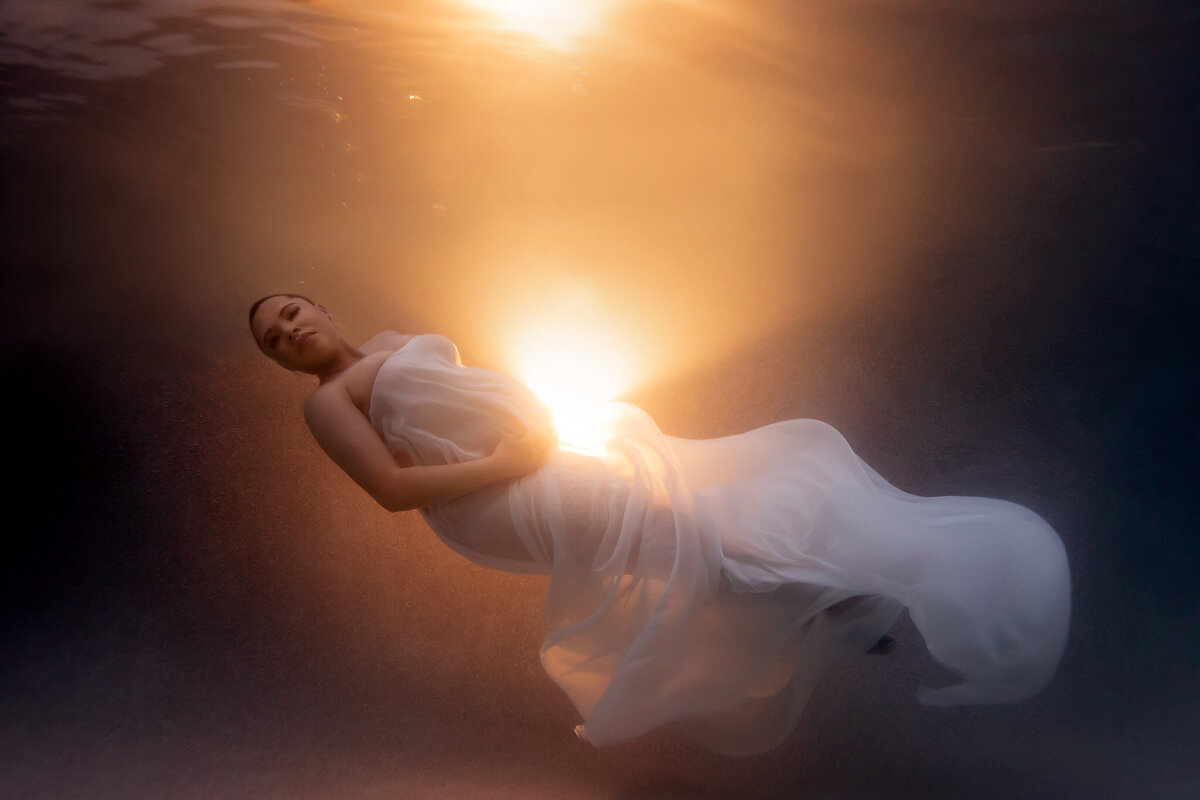 Breathtaking underwater maternity photo captured by a Dallas underwater maternity photographer. The expecting mother floats serenely in a flowing white gown that gracefully swirls around her body. A brilliant golden light radiates from behind her, creating a stunning, ethereal glow against the water. This underwater maternity photoshoot highlights both beauty and serenity in the elegant composition.