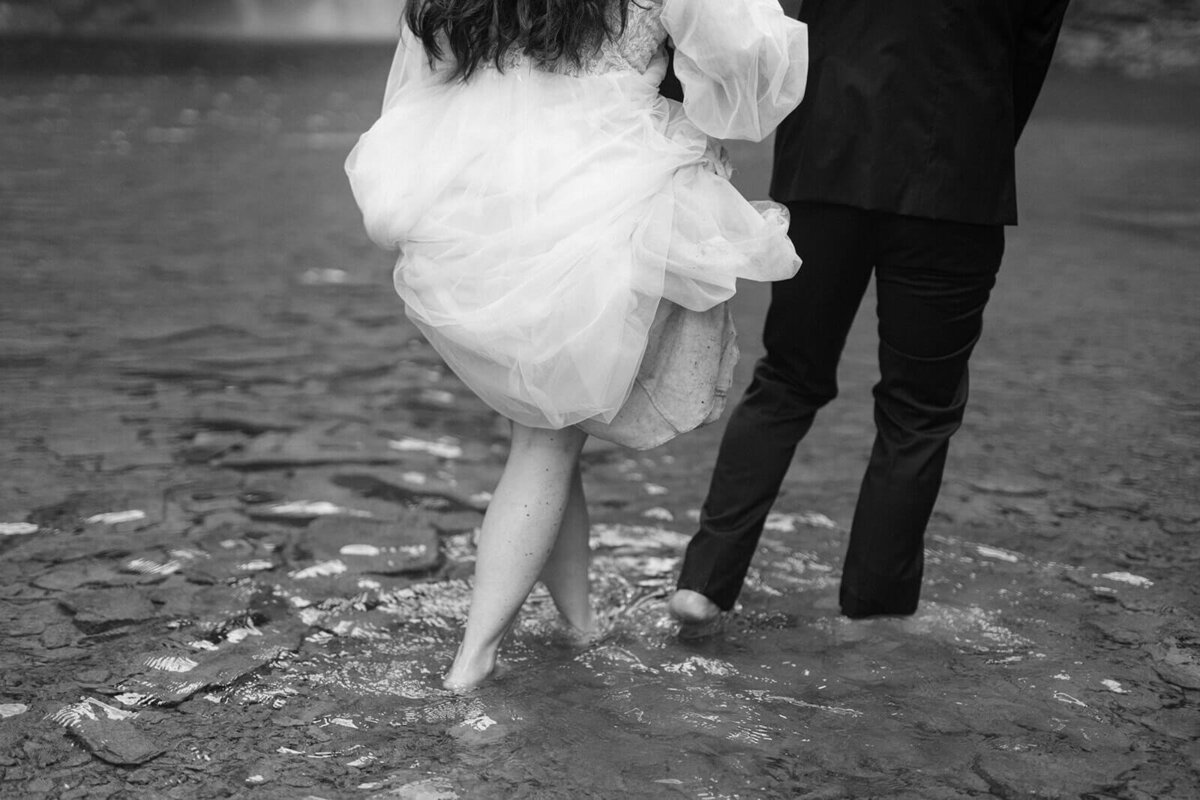 bride and groom walking in the water in front of waterfall