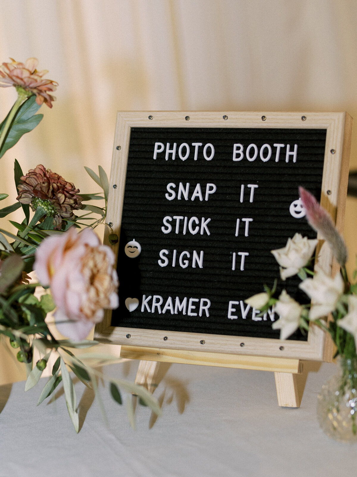 A small black felt letter board on a wooden easel surrounded by blush and cream-colored flowers. The board reads: "PHOTO BOOTH — SNAP IT  STICK IT SIGN IT KRAMER EVEN." The display is part of a romantic, floral-themed event setup.