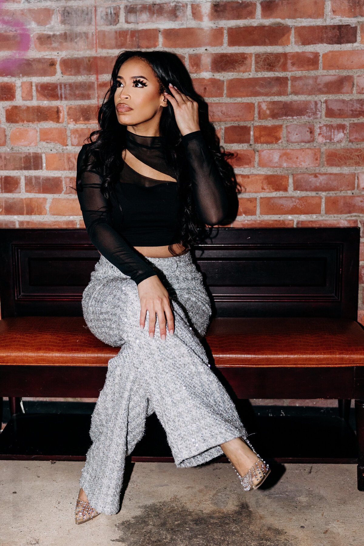 Tiara in a black cutout top and sparkly pants sitting with her legs crossed on a bench in front of a brick wall