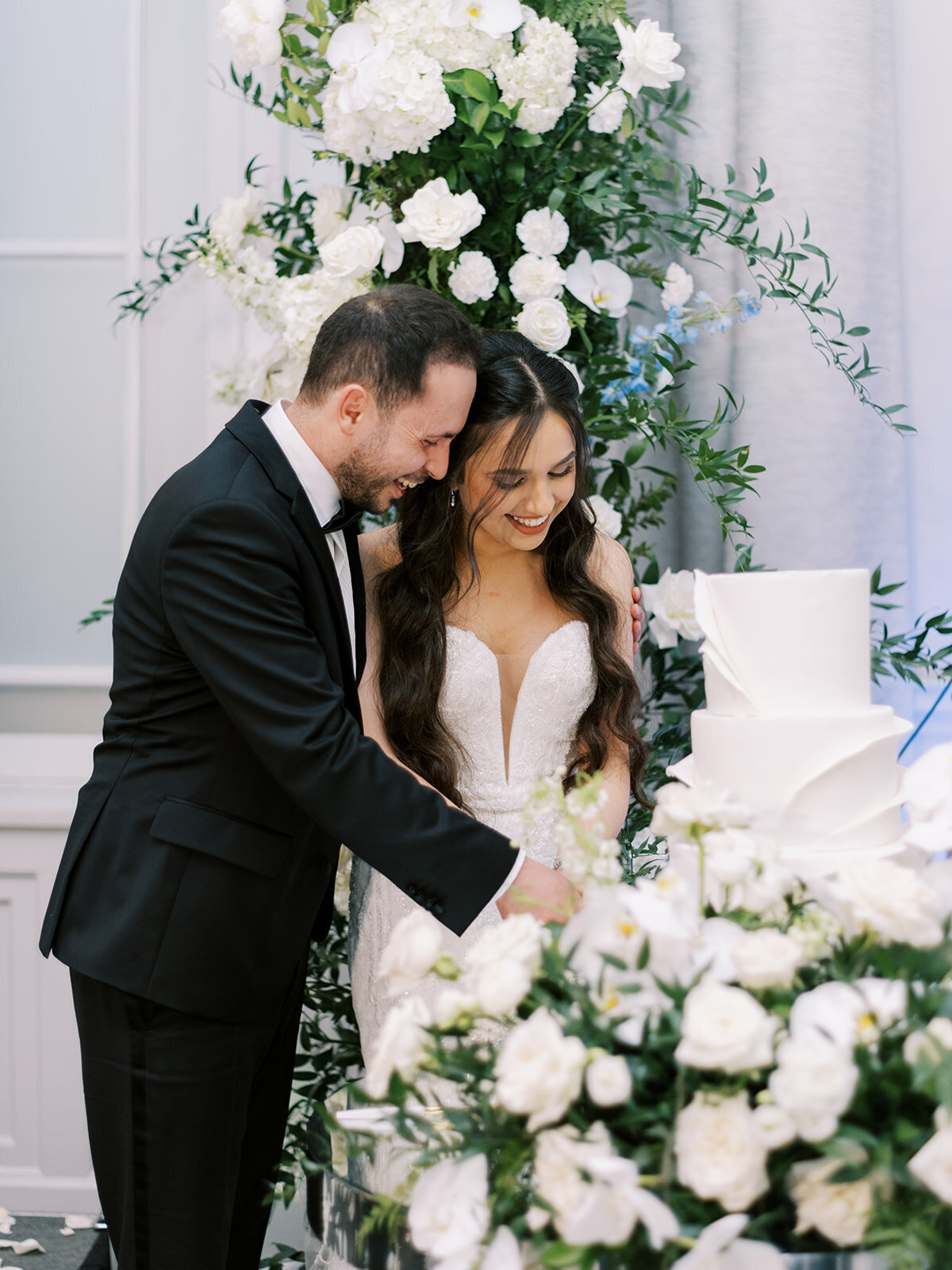 A bride and groom in formal attire smile as they cut a white, tiered wedding cake adorned with flowers, surrounded by white floral decorations at their destination wedding in Canada.