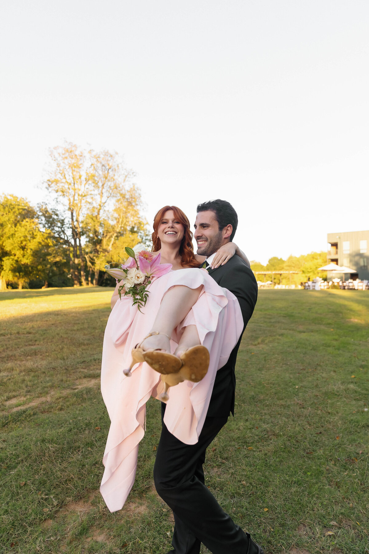 Bridesmaid in a blush pink gown laughing as she is carried across the lawn by a groomsman, holding a playful bouquet of wedding flowers designed by an Arkansas wedding florist.