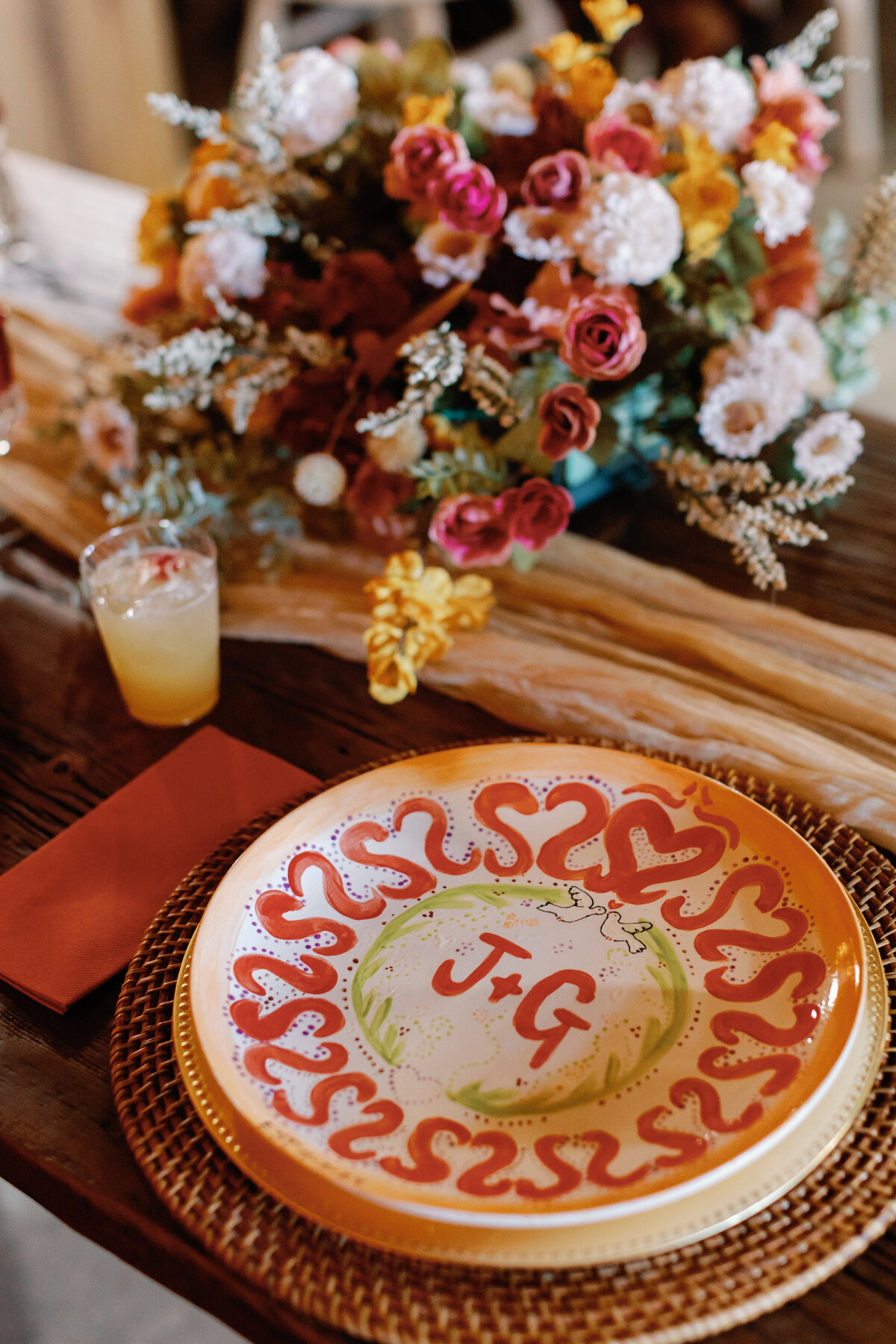 Hand-painted dinner plate with initials “J + G” surrounded by colorful flowers and cocktail on rustic wooden table.