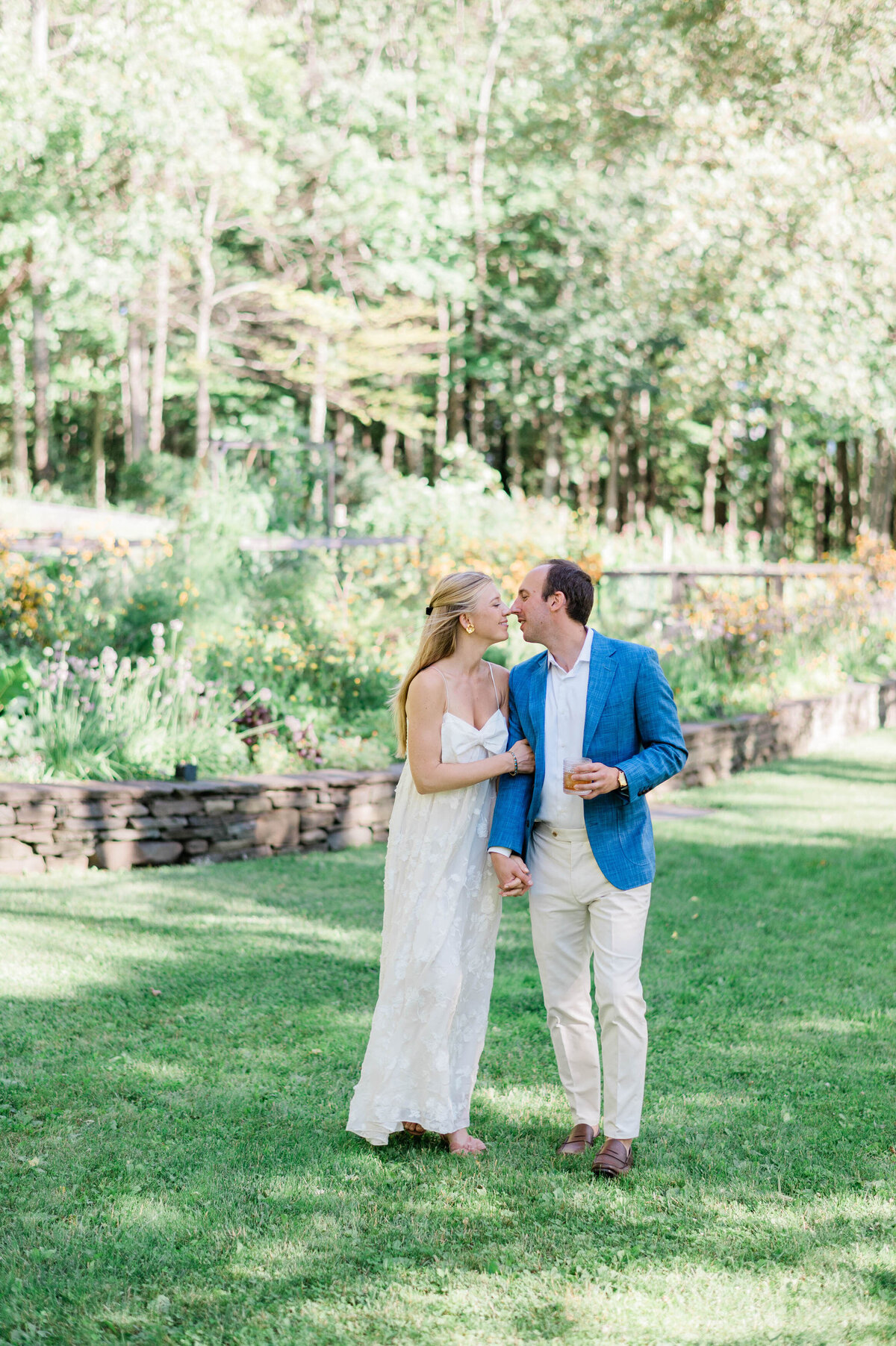 A barefoot bride in a white flowy dress and a groom in a blue blazer share a tender kiss while holding hands in a sunlit garden.