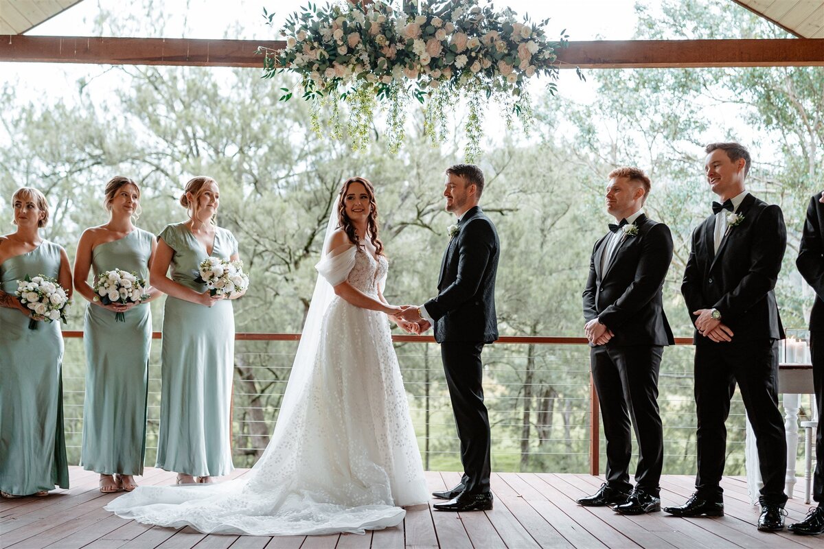 A beaming bride, clutching a bouquet of white roses, exchanges vows with her groom under a canopy of fragrant blossoms.