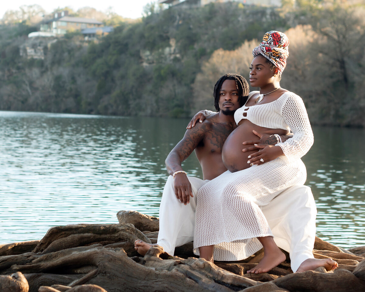 Editorial maternity portrait with a lakeside view in Austin. The couple sits on sculptural tree roots with the expecting mother in a white boho gown.