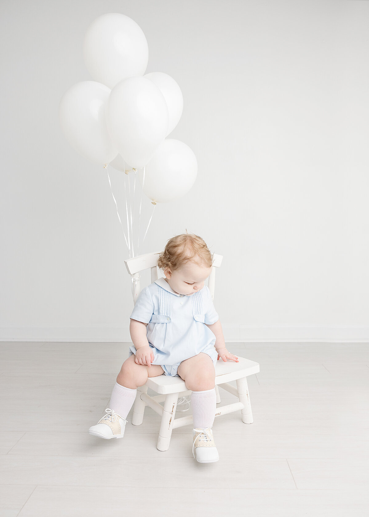baby boy photographed in studio in a feltman brothers outfit with white balloons on a white chair