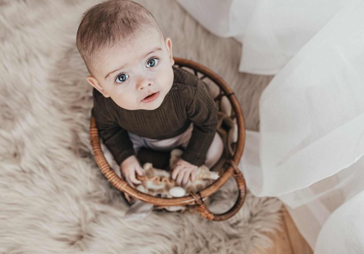 Ruua Hamdan Photography (UK): A baby with wide eyes sits in a wicker basket on a furry rug.