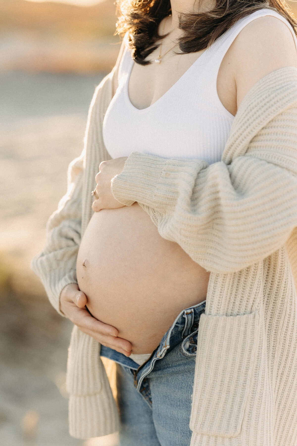 Mother-to-be in casual maternity outfit holding her baby bump at Coronado Beach during sunset. Captured by San Diego Family Photographer Yuka Schad.