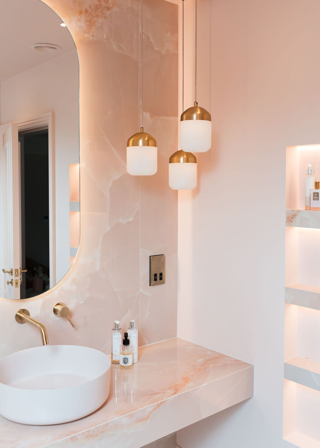 A modern pink marble vanity featuring a white sink bowl with gold hardware. There is a chic oval mirror over the sink and a trio of white and gold pendant lights hanging in the corner.