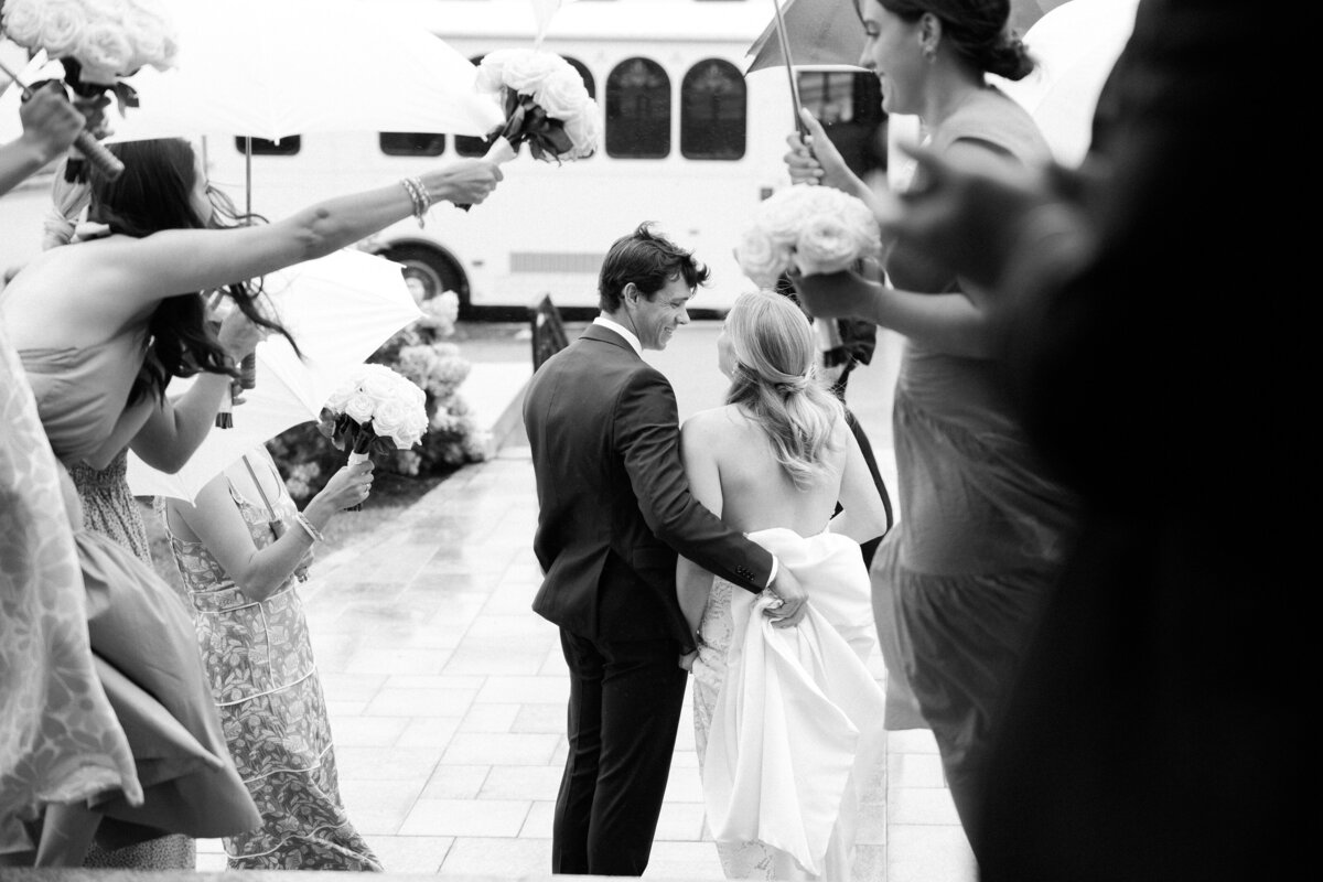 the bride and groom walking up the steps of Spring Lake Golf Club, surrounded by friends holding white umbrellas and bouquets of white roses. The groom smiles at the bride as he lifts the train of her dress.