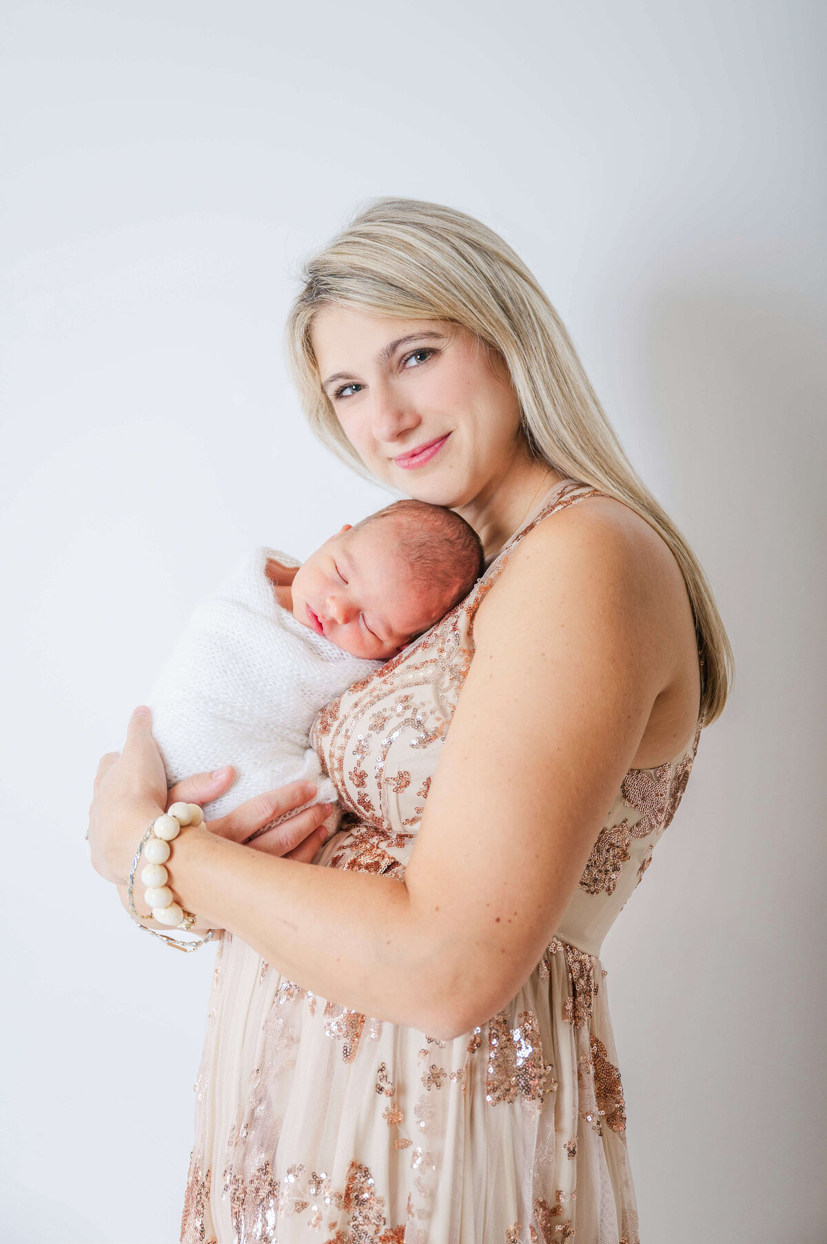 A mother in a sequined dress smiling at the camera while holding her newborn baby on a white backdrop.