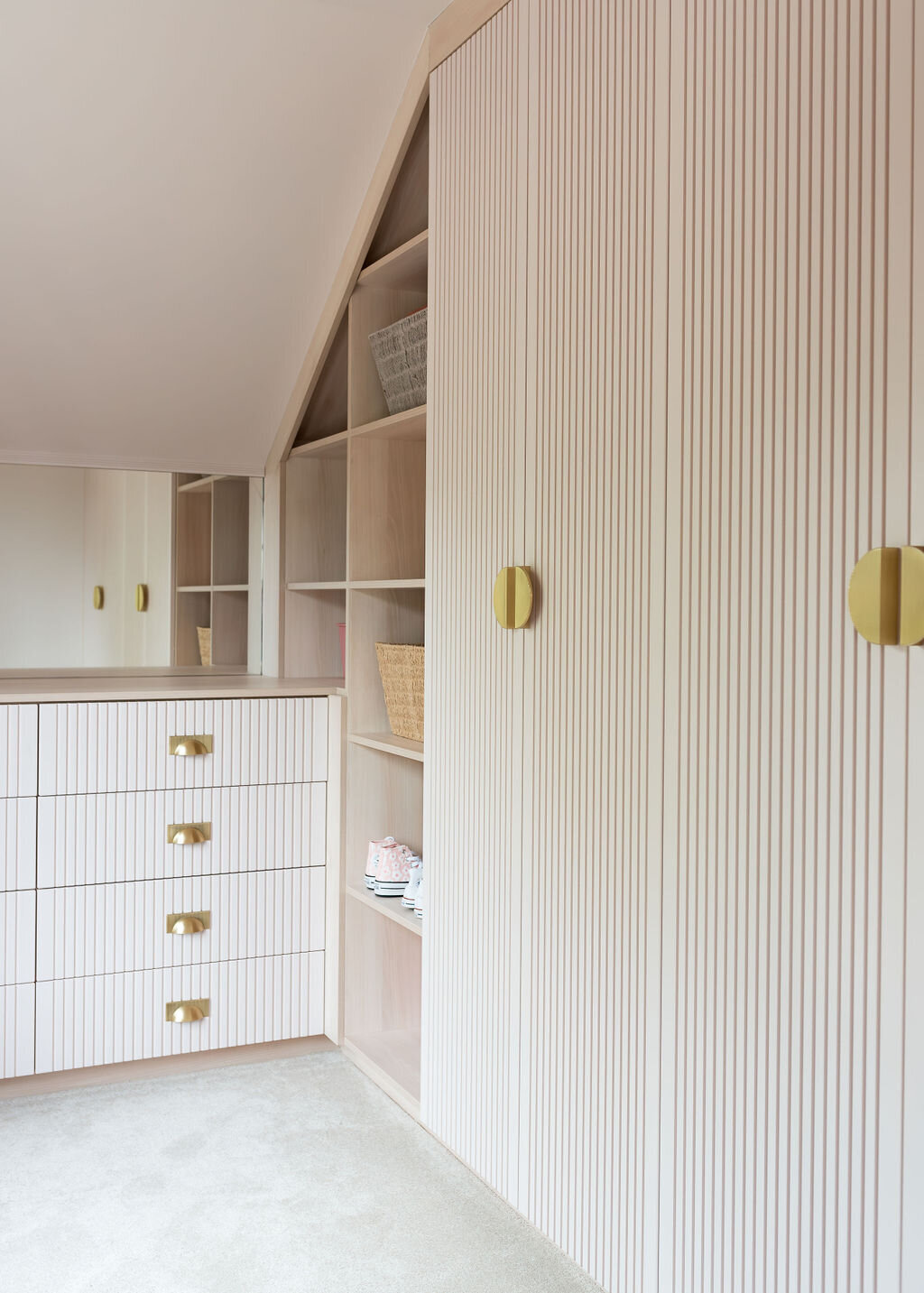 A modern pale pink wardrobe with vertically striped doors and large circular gold handles. To the left, a modern pale pink dresser with gold half-moon handles. The dresser has a ribbed texture on the drawer fronts and features four drawers.