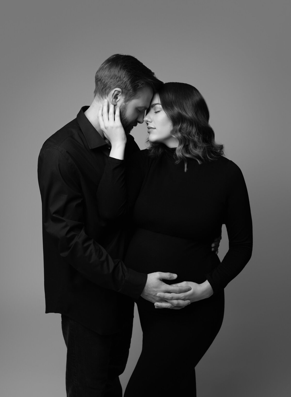 Emotional studio maternity photo in black and white of a couple embracing with foreheads touching, dressed in all black.