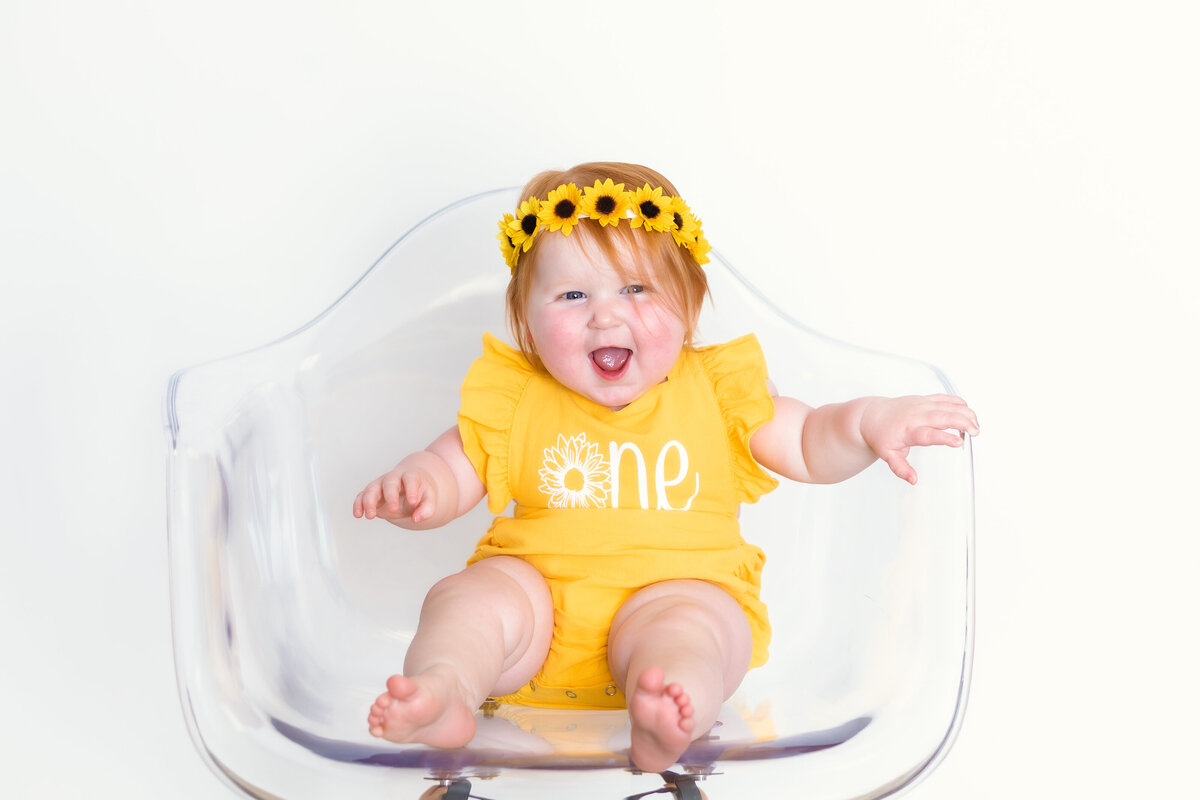 Cake Smash Photographer, a baby girl sits on a chair with a onesie that reads "one" and wears a headband that has sunflowers