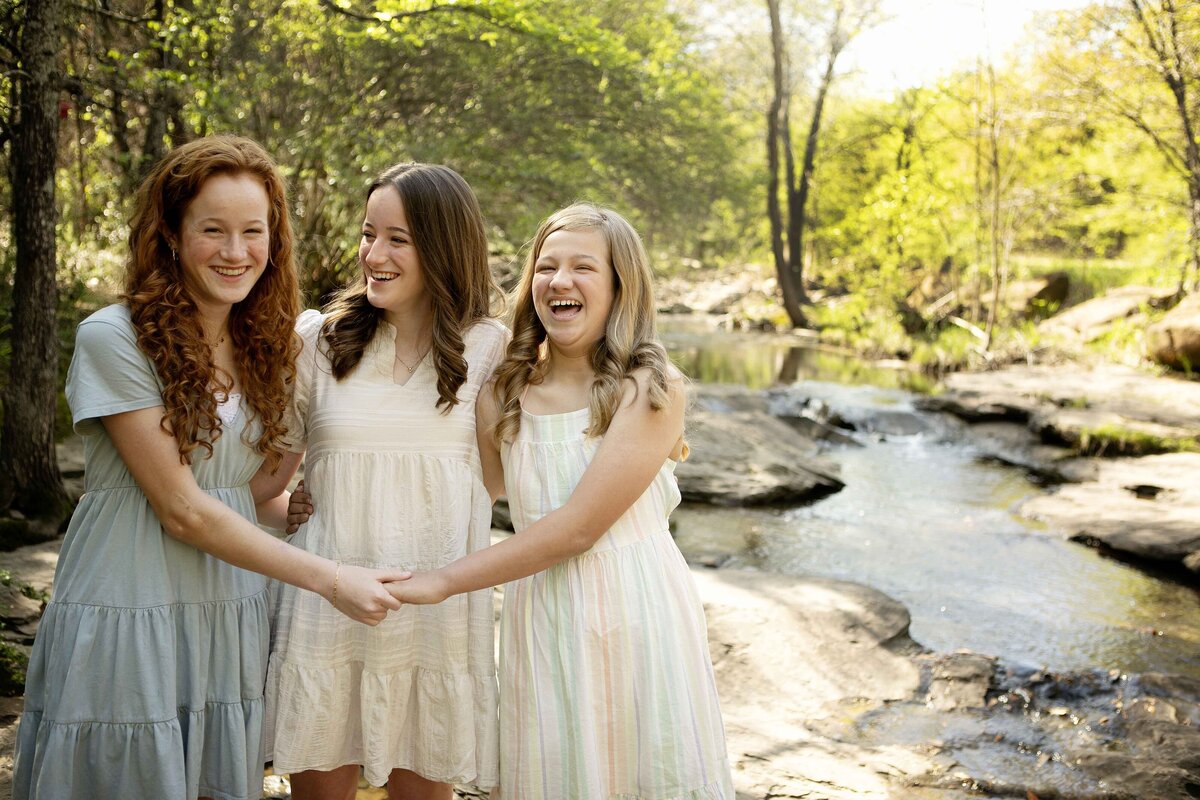 Three sisters standing by the creek in Flower Mound, their dresses flowing in the breeze as they pose for a picture