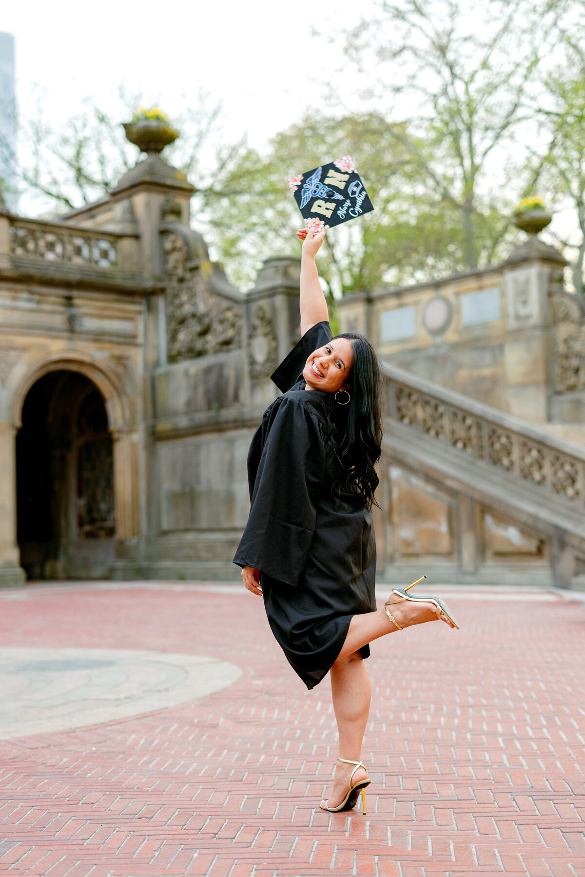 Hostos Community College graduate smiling while holding cap in the air at Central Park.