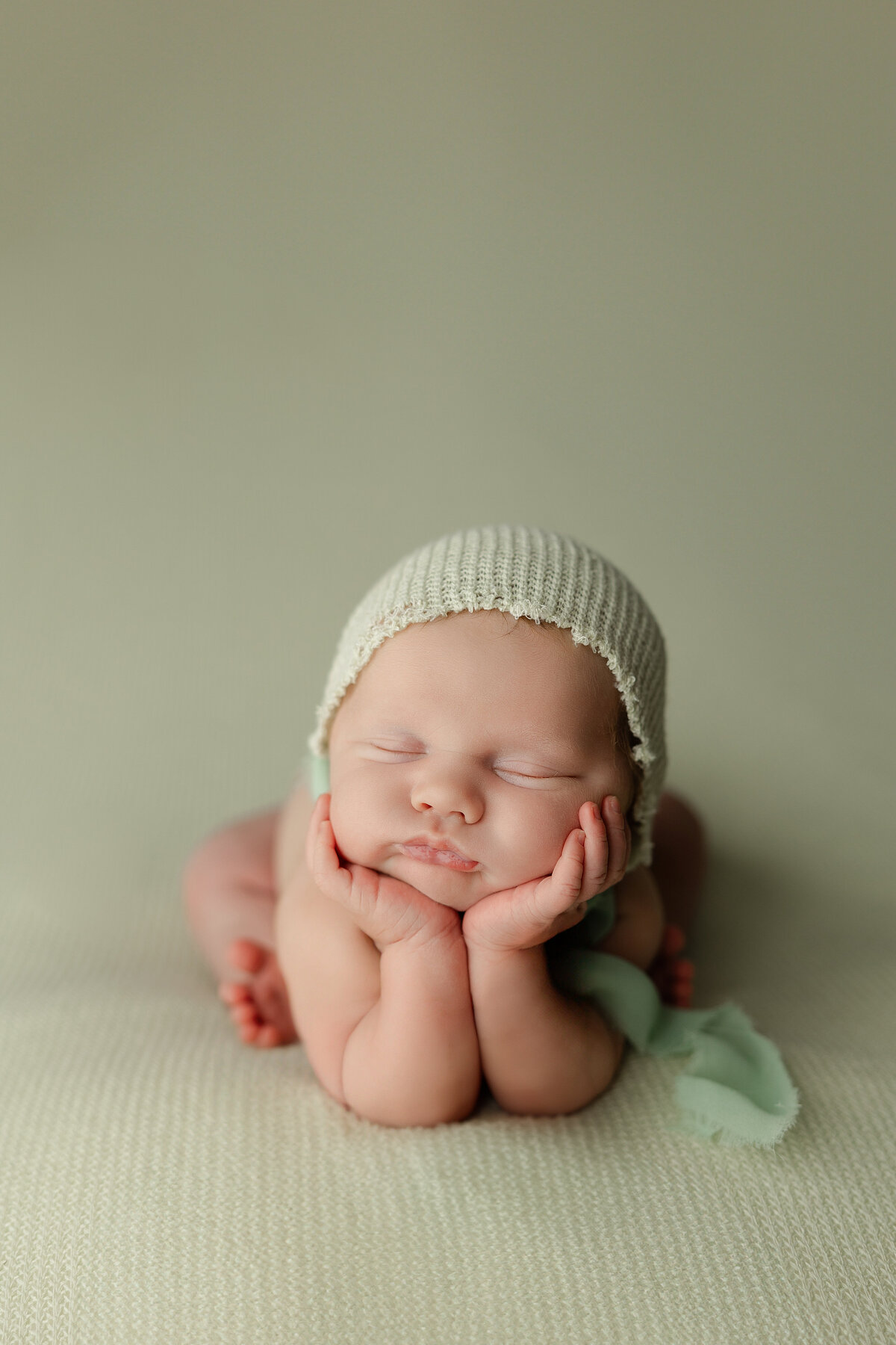 Newborn baby posed in the classic froggy position with a cream knit bonnet on a soft green backdrop during their studio session. Captured by Inland Empire newborn photographer Kaitlyn Dawn Photography.