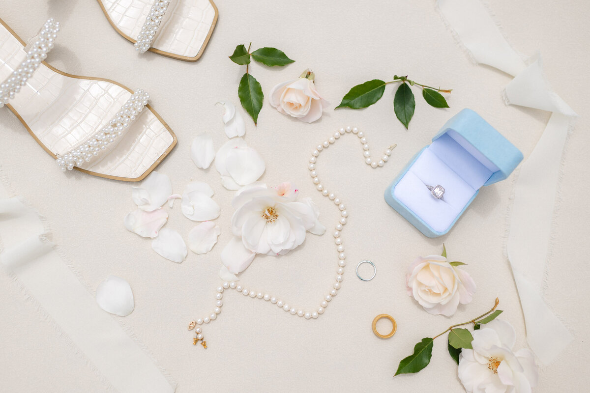Flat lay of bridal accessories with pearl sandals, engagement ring in a blue box, pearl necklace, and white roses