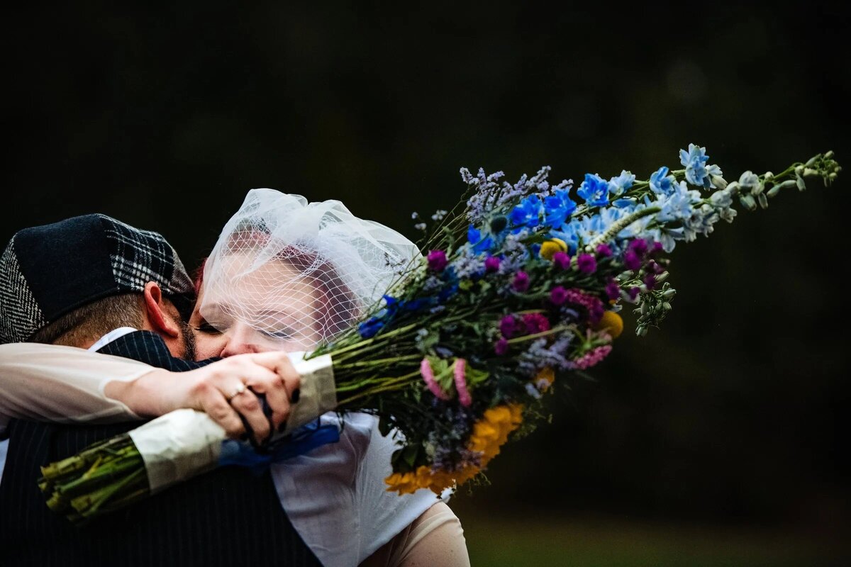 A romantic close-up of a bride in a veil and groom in a cap, kissing behind a vibrant bouquet, focusing on the bouquet in the foreground.