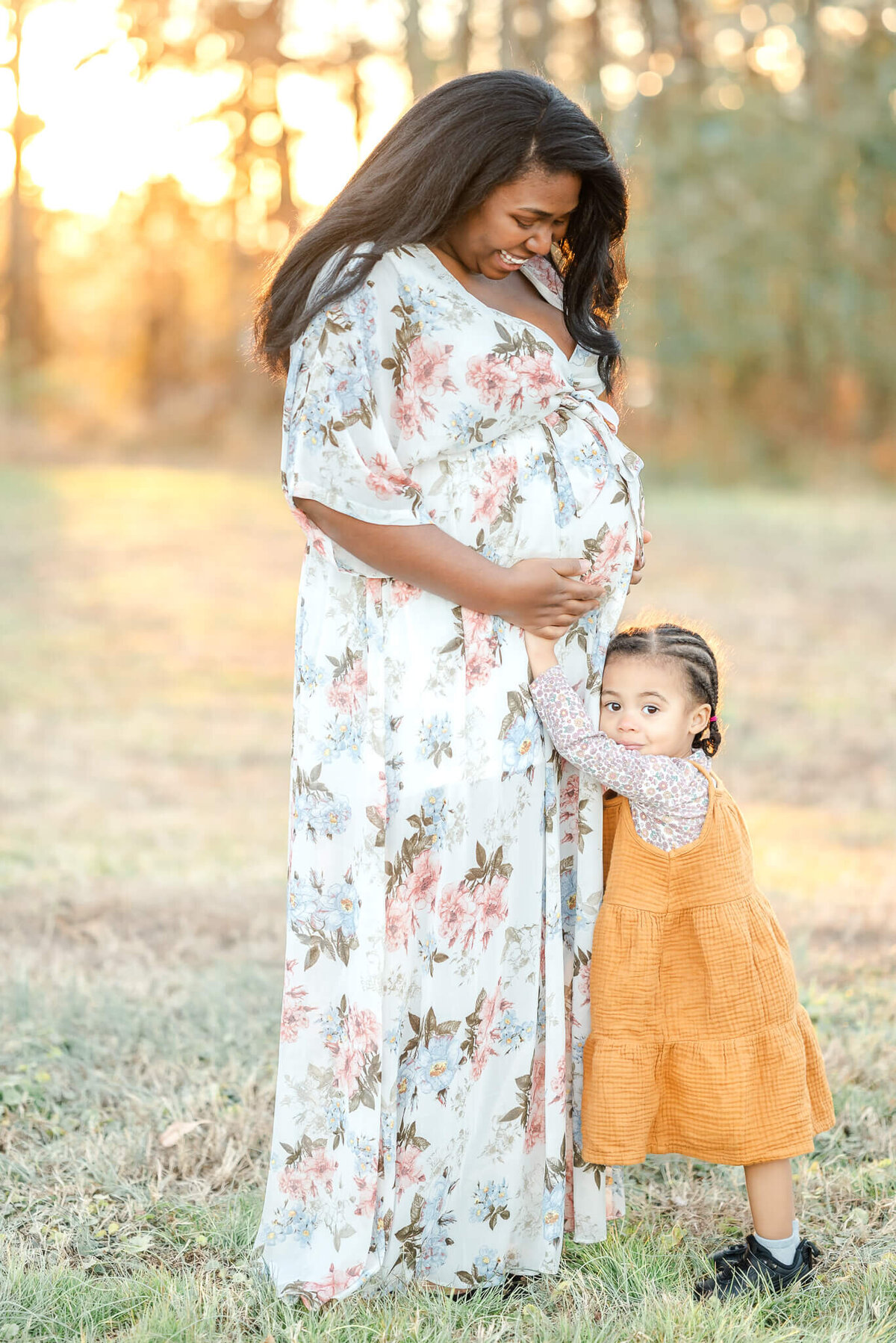 A toddler, wearing a mustard color dress, holds onto her mama's belly. Mama, wearing a white floral, smiles down at her.