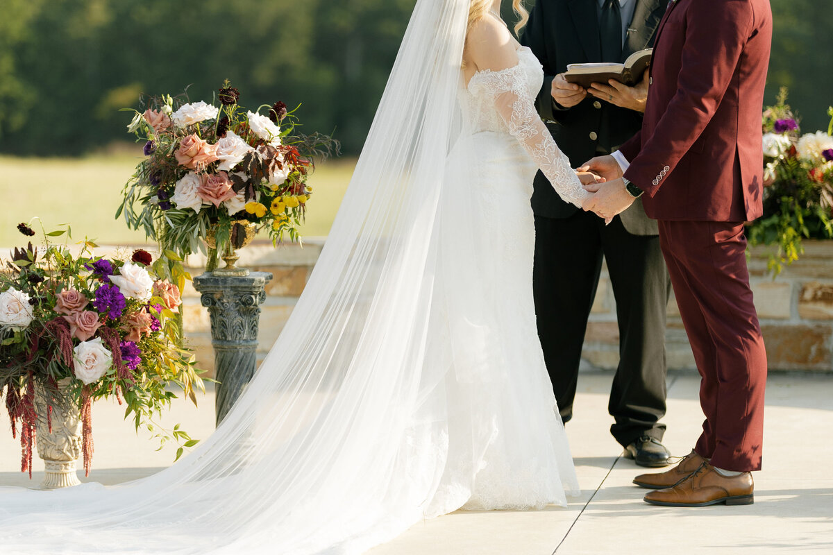 Bride and groom holding hands during their outdoor ceremony framed by lush mauve, blush, burgundy, and purple floral arrangements designed in an organic, garden-inspired style by a wedding florist specializing in romantic luxury weddings.
