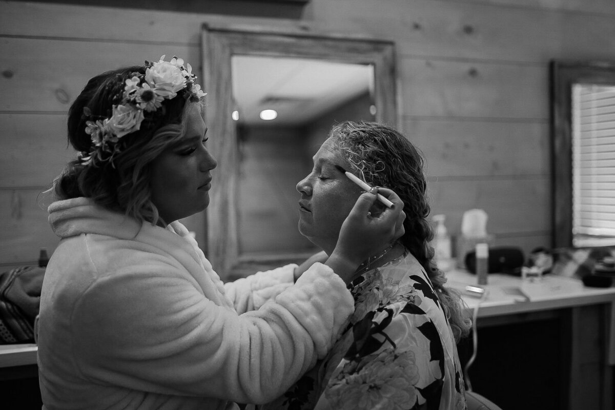 Bride applying makeup to a seated woman adorned with a floral crown