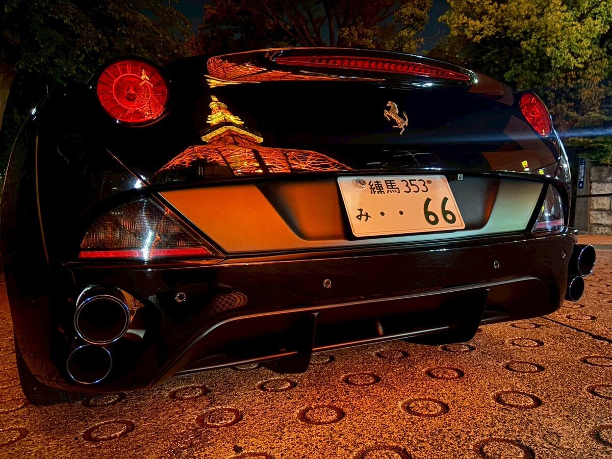 Rear view of a black Ferrari parked at night, with Tokyo Tower’s warm lights dramatically reflected on its sleek surface
