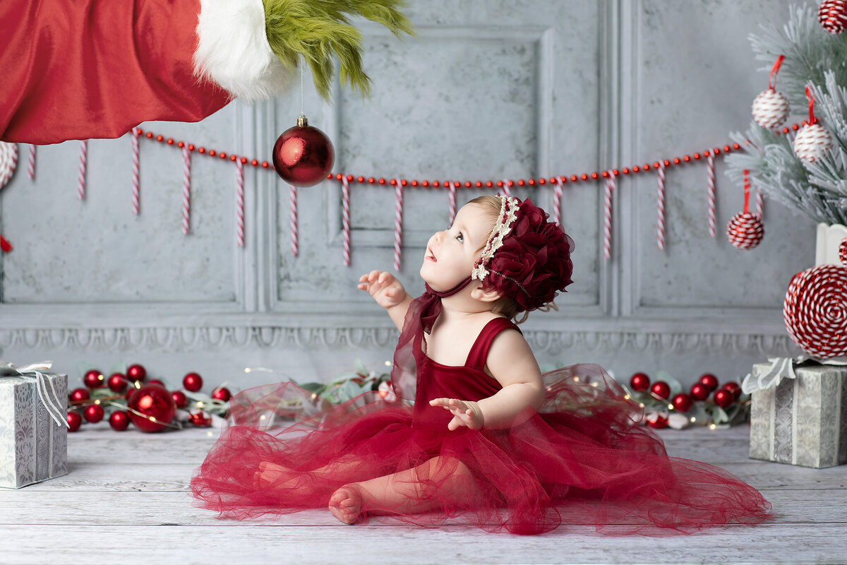 Baby girl in a festive red dress gazes up at a red ornament held by a furry green Grinch hand during a whimsical Dallas milestone photographer holiday session.