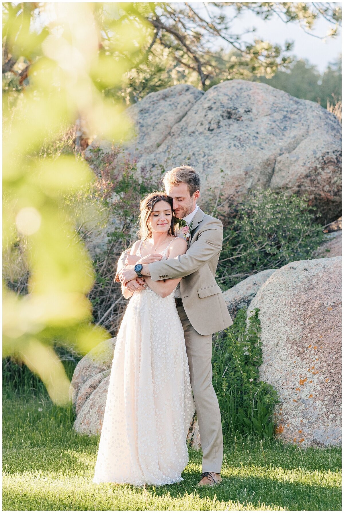 A groom in a tan suit hugs his new bride from behind as they both stand in front of a large boulder