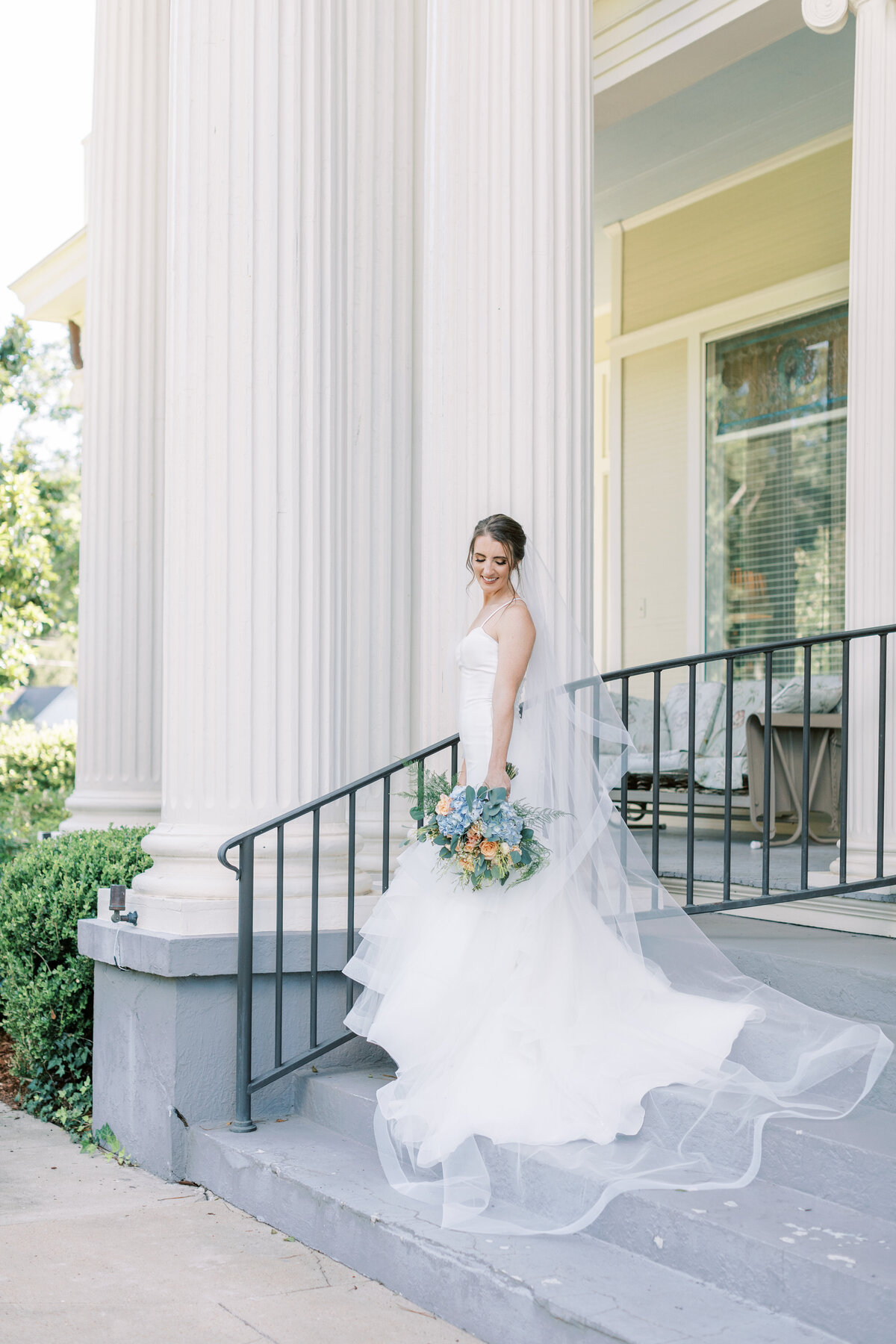 A bride looks down at her bouquet while standing by a white column.