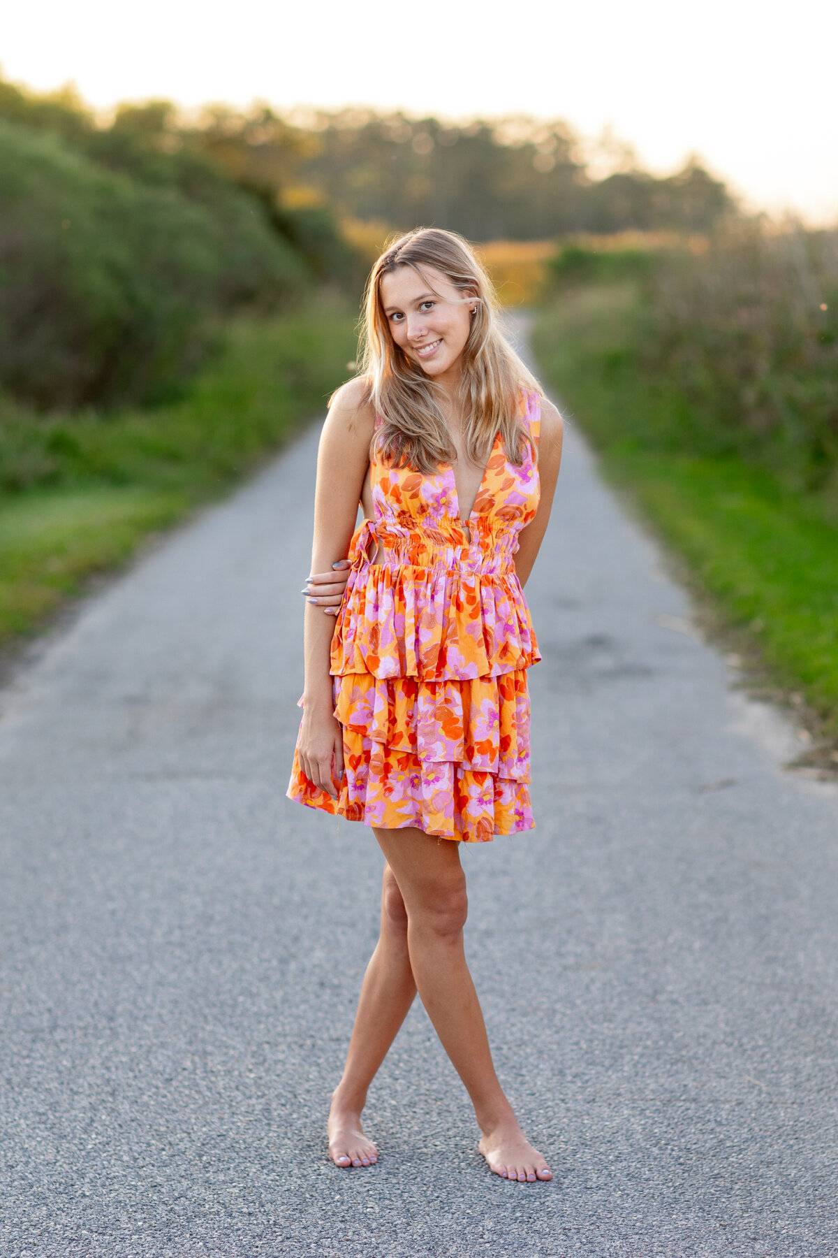 A high school senior in a vibrant floral sundress stands barefoot on a quiet country road, smiling with relaxed confidence.  Captured by Brooke Tucker Photography in rural Virginia, this portrait blends playful style with timeless Southern Charm.  