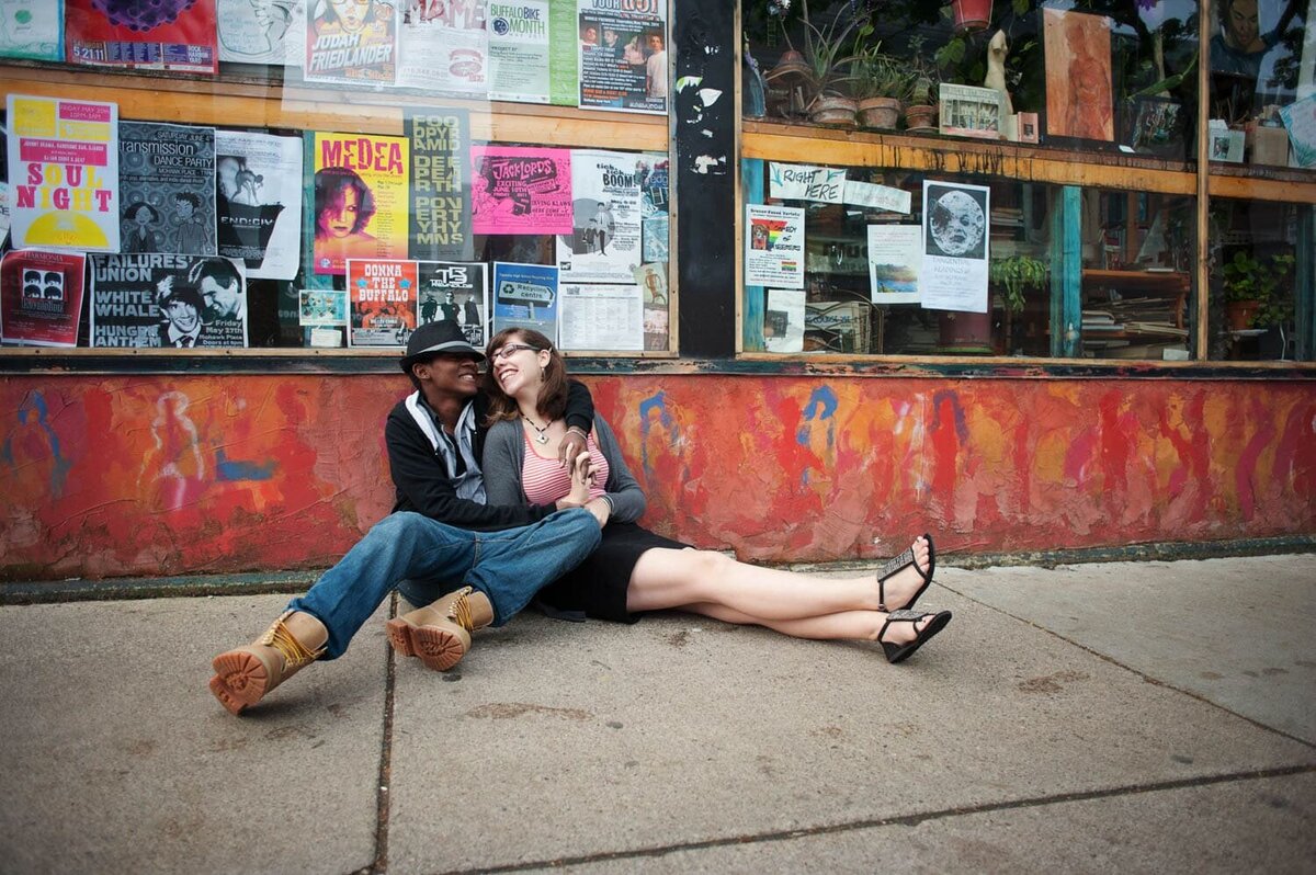 a fun couple hugs together while sitting on the ground in front of a music venue with a colorful mural wall