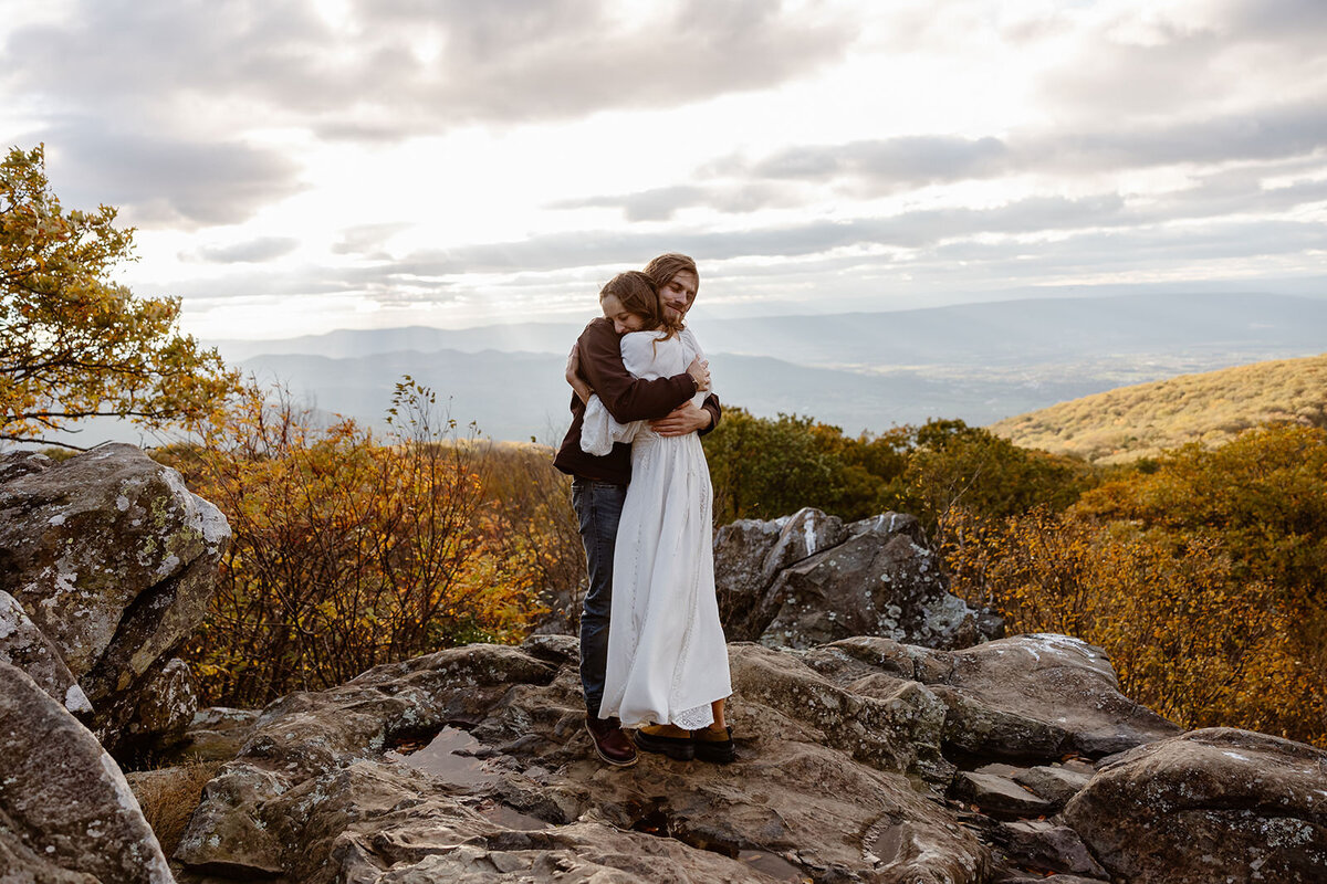 A couple embraces as they stand on Hawksbill Summit. There is a huge gust of wind, and it maybe them embrace even tighter to stay warm.