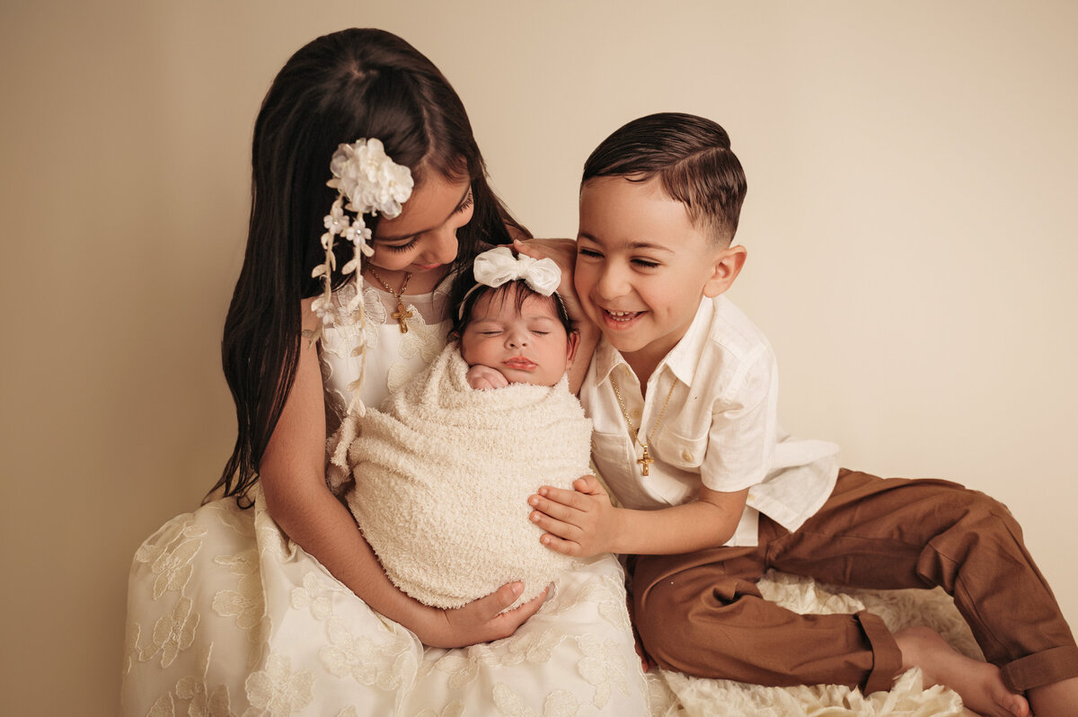 brother and sister siblings wearing beige holding their newborn baby sister who is swaddled in a cream wrap and is sleeping