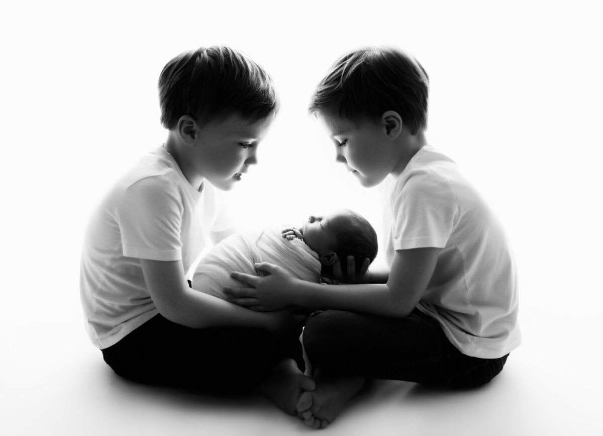 Young brothers sitting cross legged facing each other and tenderly looking at their newborn sibling wrapped in the middle of them.