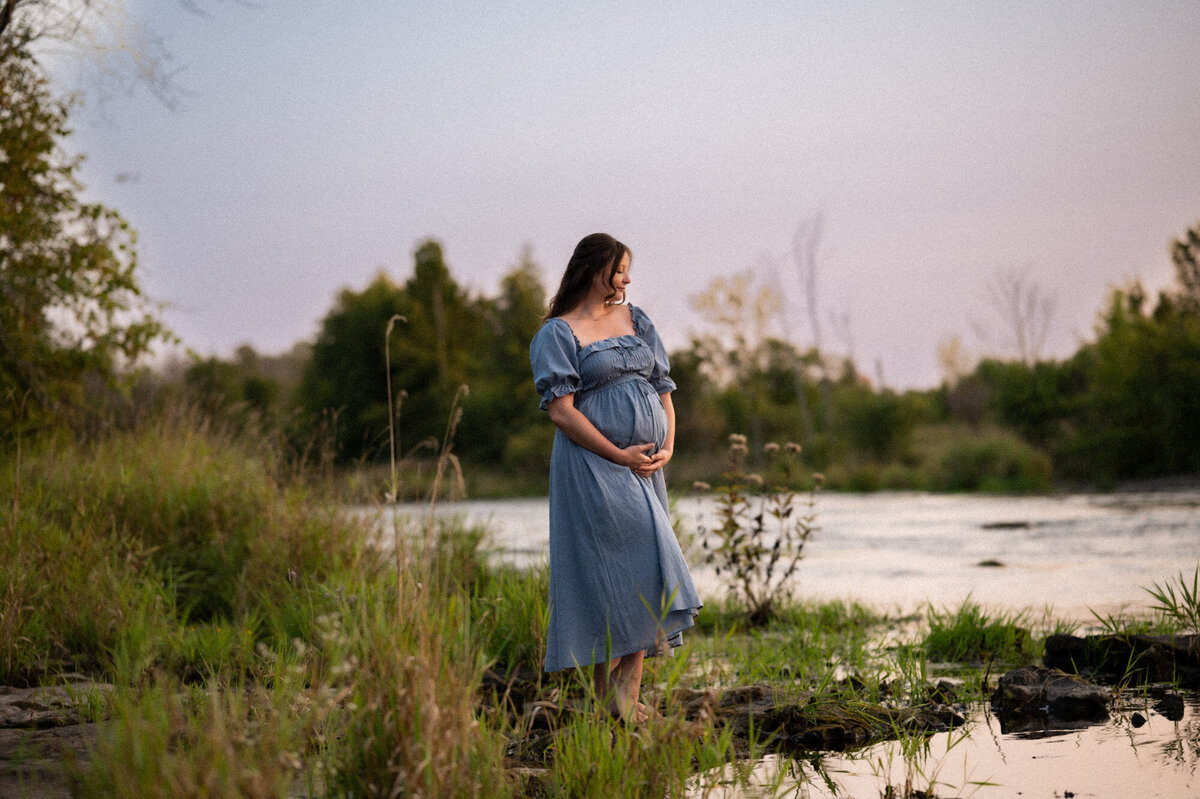 Woman holding her pregnant belly, wearing a blue maternity dress by the Jock RIver.