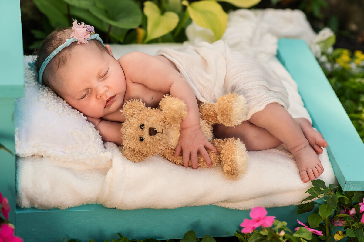 Infant girl sleeping on a blue bed on a bed of flowers outdoors holding a teddy.