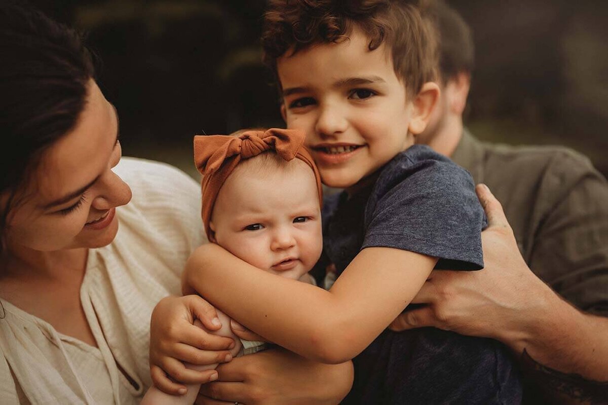 a mom holds her baby girl and the dad holds his son as as the son embraces his younger sister in a gentle hug